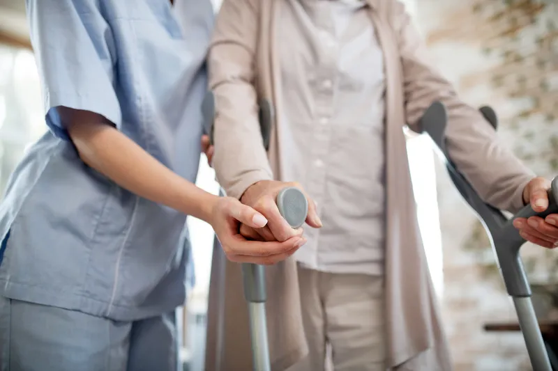 Professionelle Unterstützung im eigenen Zuhause. Eine Caritas-Pflegekraft in blauer Arbeitskleidung unterstützt eine Seniorin beim Gehen – Ambulante Pflege im Raum Teublitz.