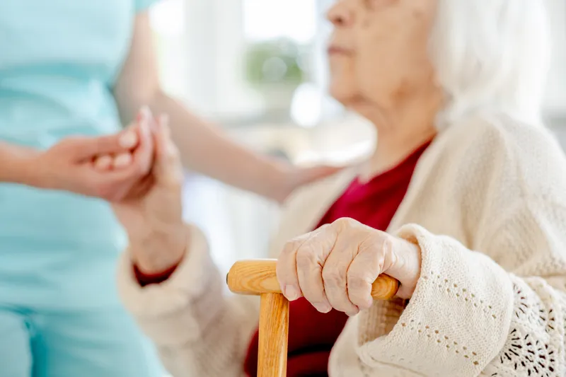 Hilfe zur Selbstständigkeit. Eine Pflegekraft stützt die Hand einer Seniorin am Gehstock – Unterstützung zur Erhaltung der Mobilität im Alltag.